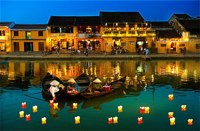 Na foto vemos três pessoas, cada uma num barco, vestindo trajes tipicamente asiáticos, colocando lanternas flutuantes sob o rio de Hôi An 